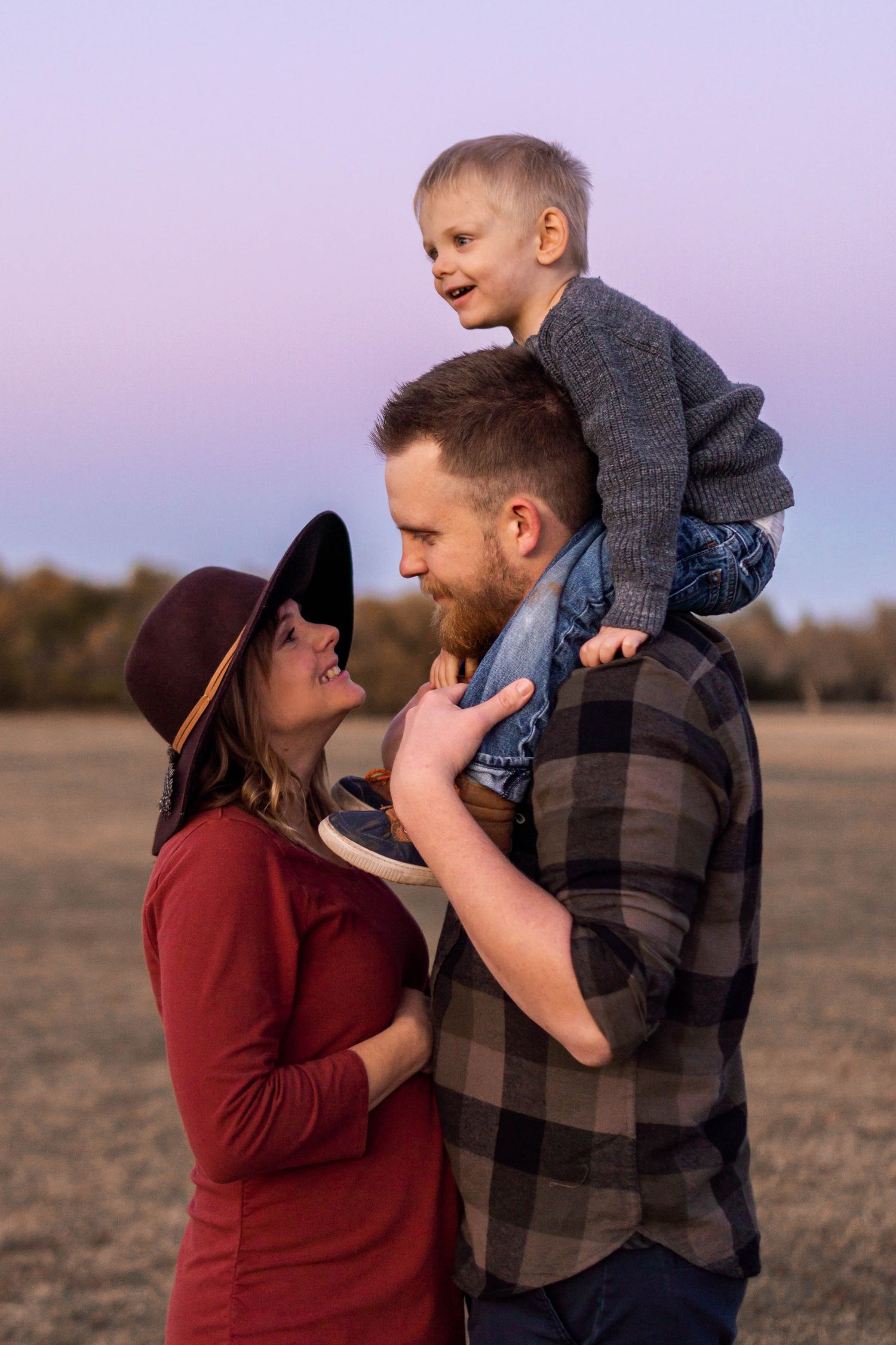 A happy family poses at sunset with dad carrying their laughing toddler on his shoulders while mom smiles beside them.