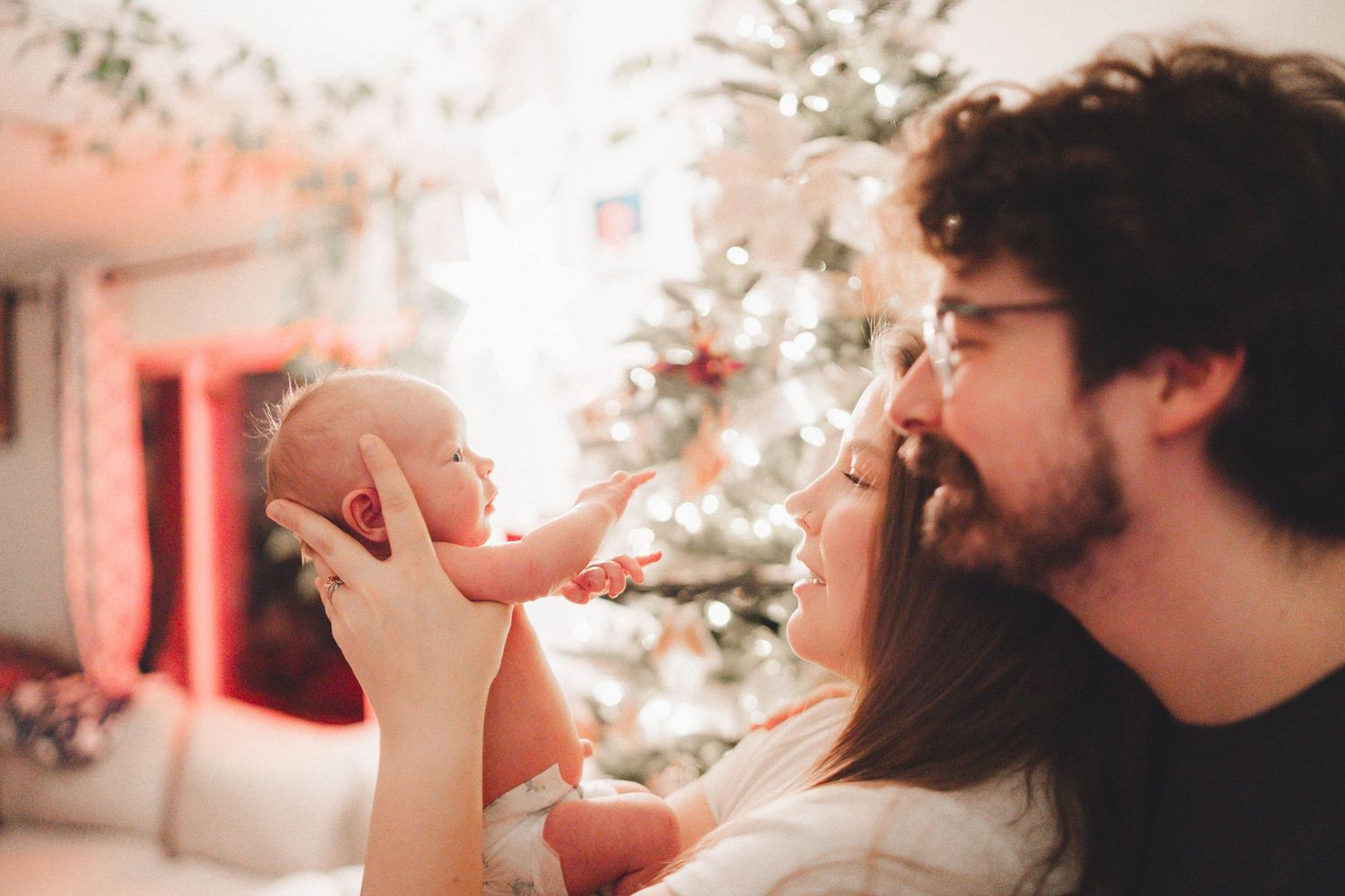 Young couple holding newborn baby near decorated Christmas tree, sharing a tender family moment.