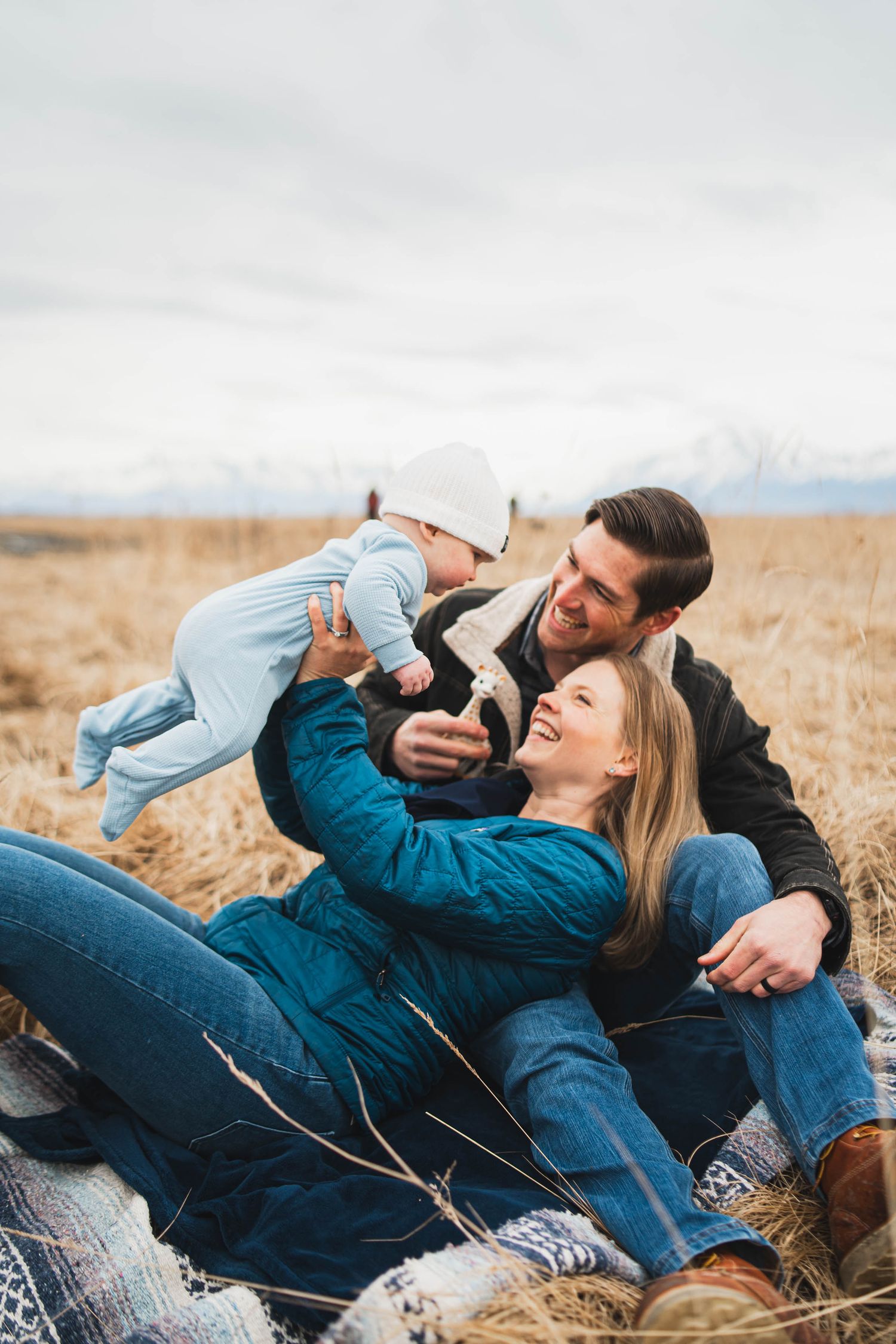 Happy young family sitting in golden field, parents lifting baby in blue onesie, snowy mountains backdrop.