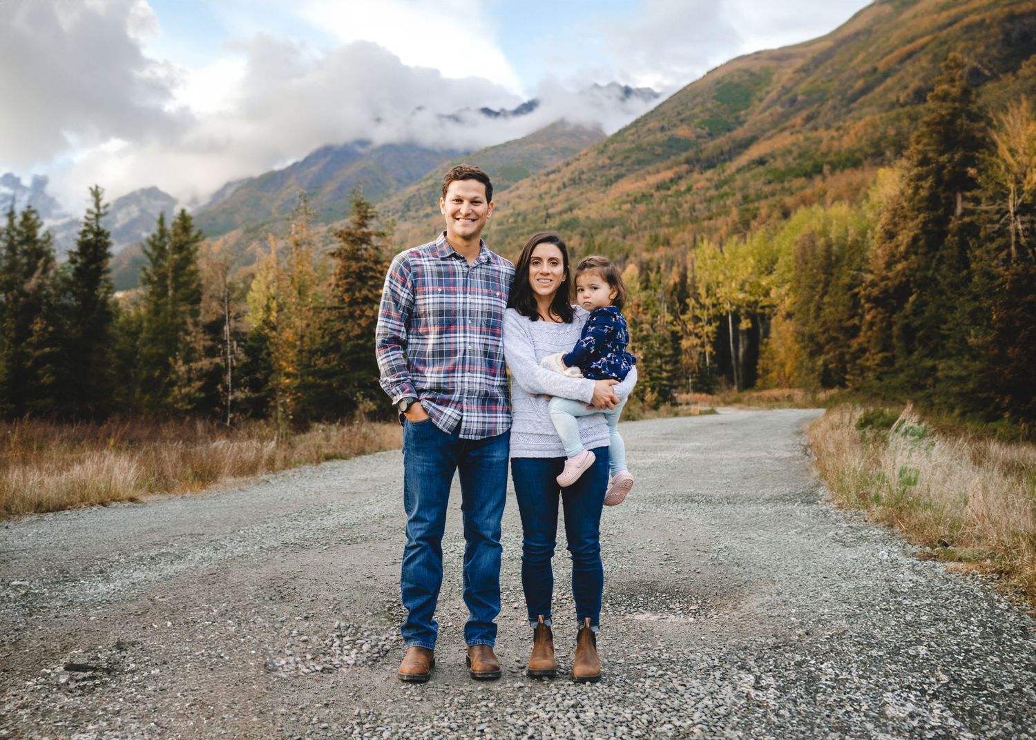 A happy young family of three poses on a gravel road with colorful autumn mountains behind them.