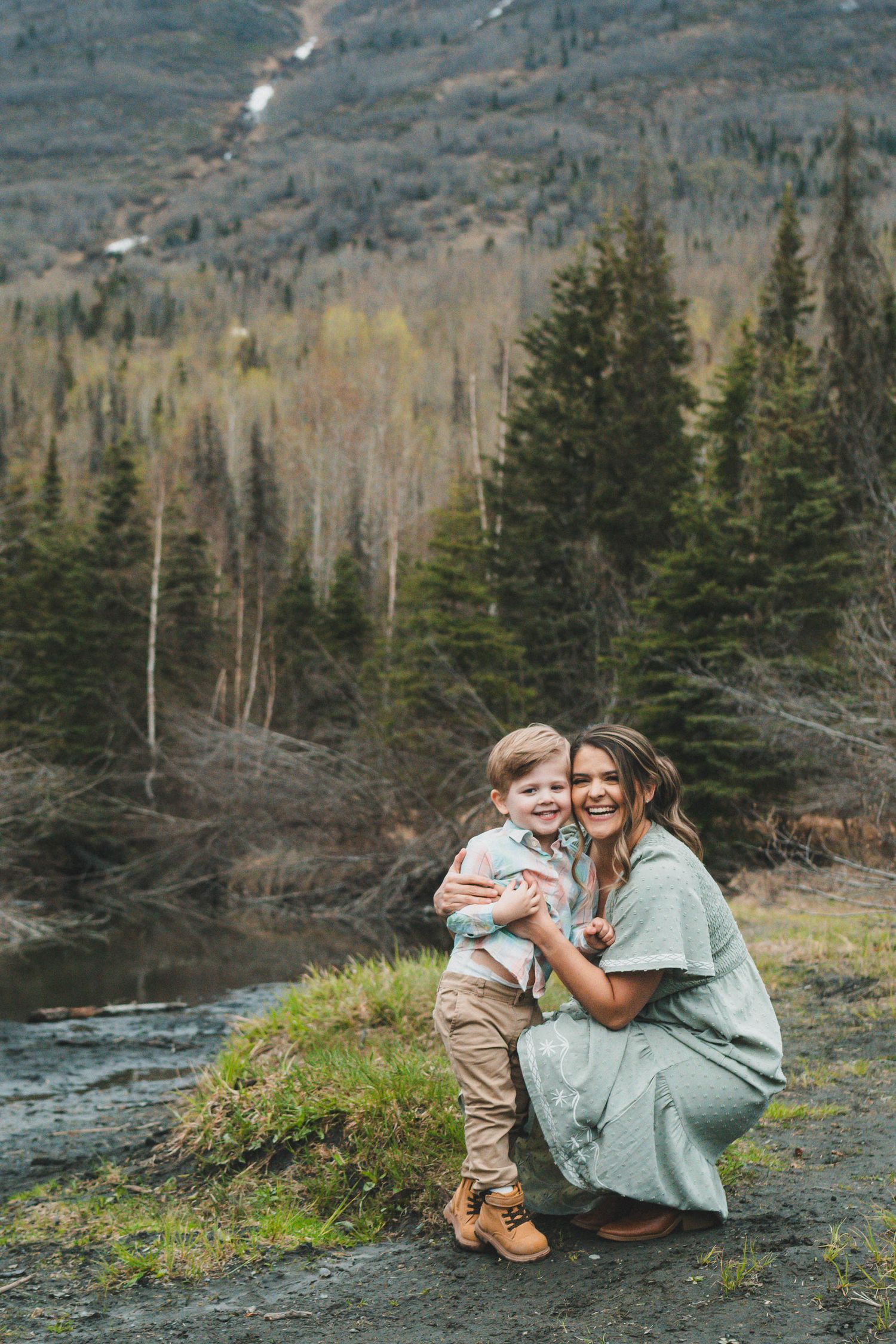 Smiling mother in a mint dress hugs her young son near a forest creek in a spring mountain setting.
