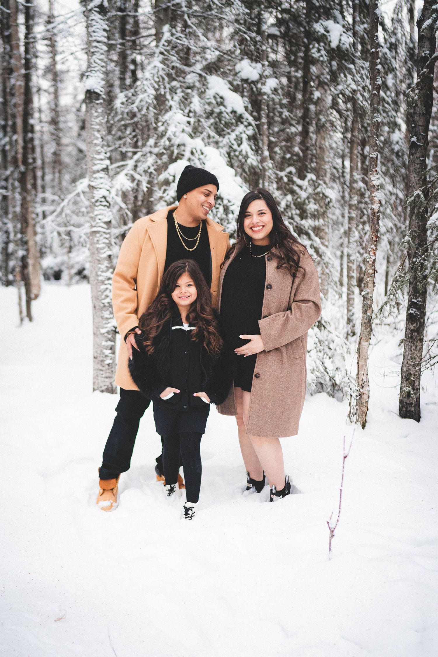 Family of three posing in snowy winter forest, parents in tan coats with child, maternity photo session outdoors.
