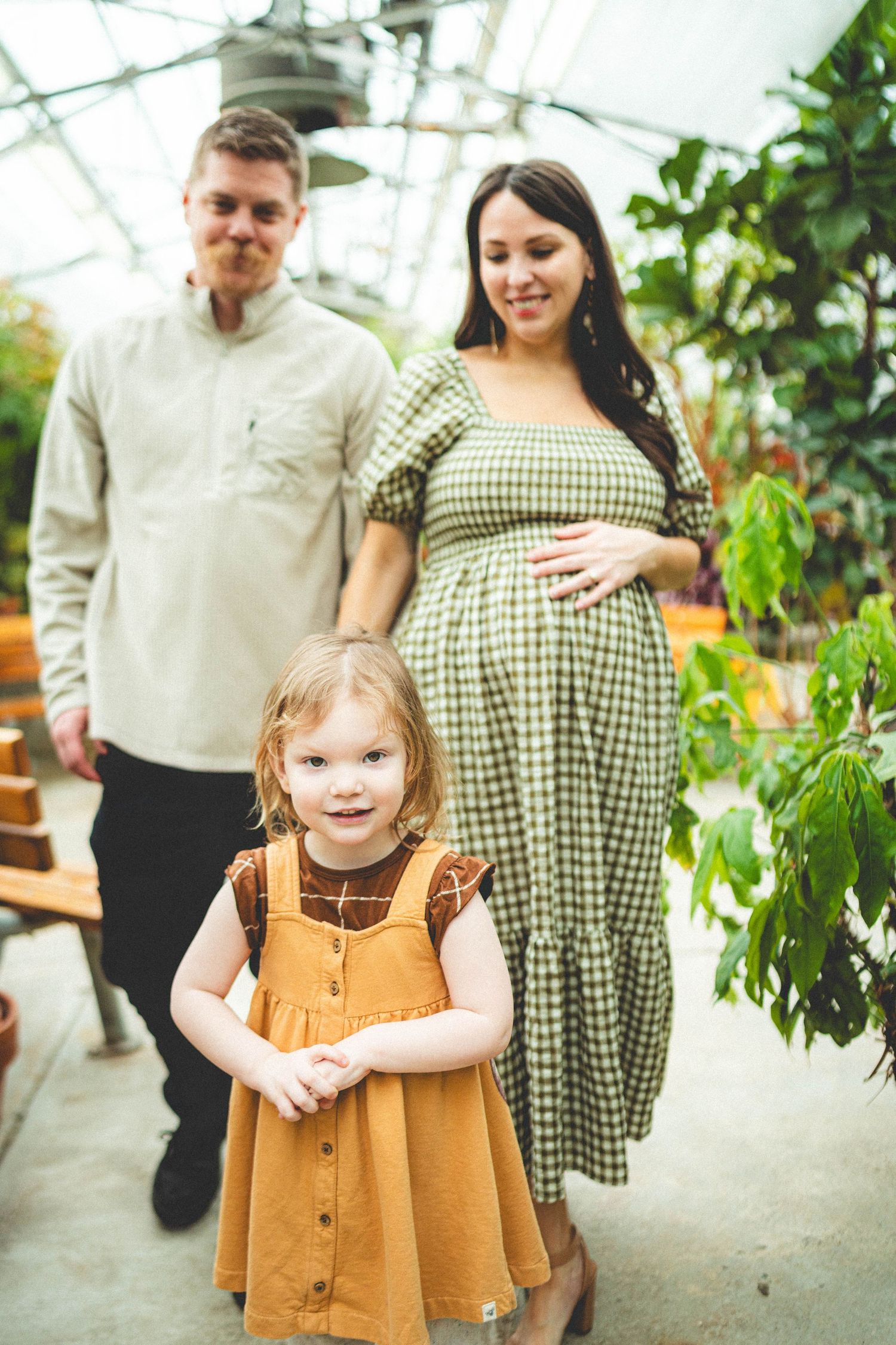 A pregnant woman in a green dress with her partner and young daughter in an orange dress inside a greenhouse.
