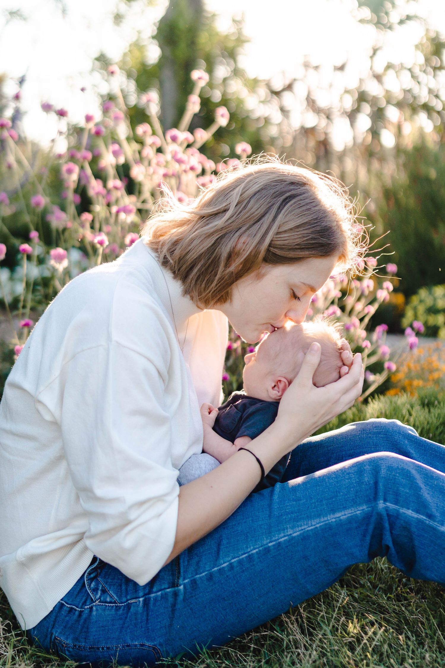 A mother tenderly kisses her newborn baby while sitting in a sunlit garden with pink wildflowers.