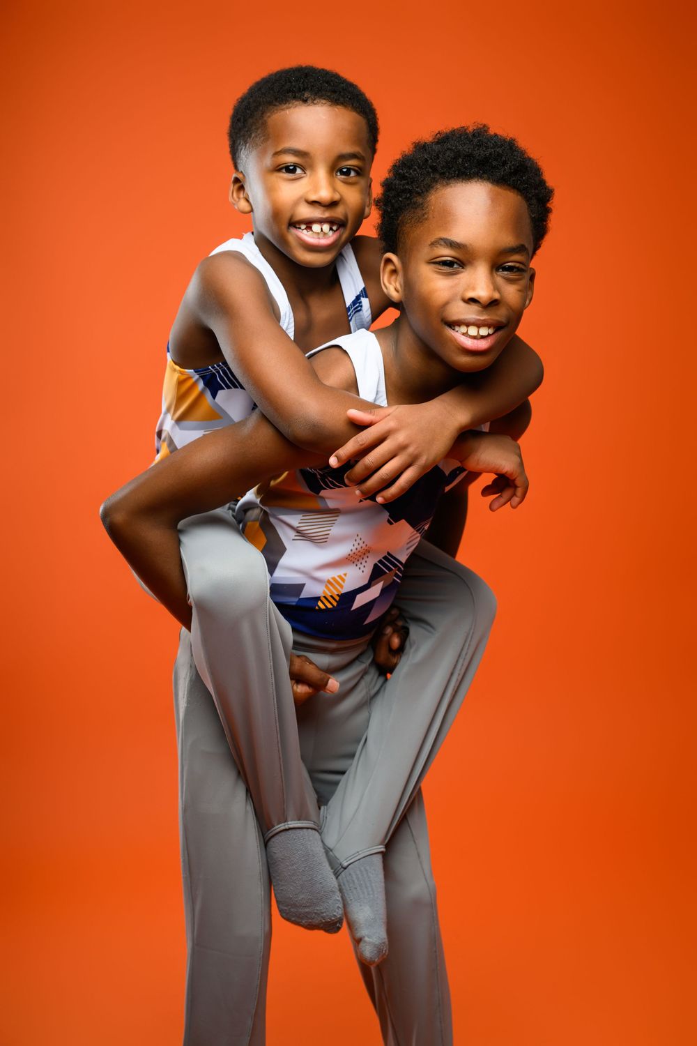 Two young African American boys smiling, one giving the other a piggyback ride against an orange background.