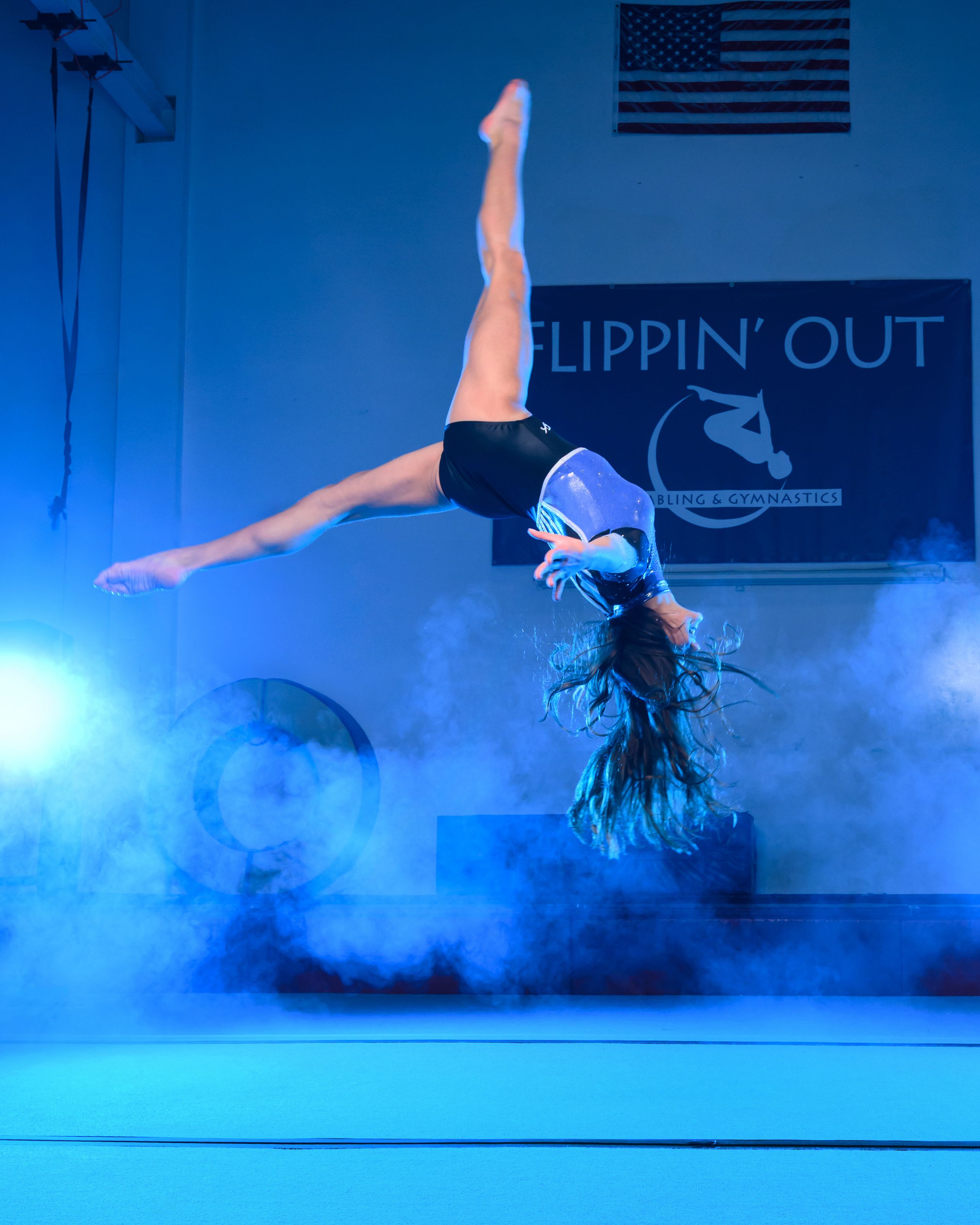Dancer performing a dynamic handstand in blue atmospheric fog on stage with an American flag in the background.