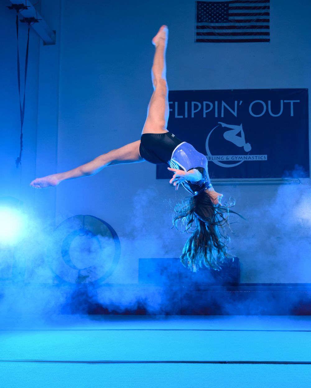 Dancer performing a dynamic handstand in blue atmospheric fog on stage with an American flag in the background.