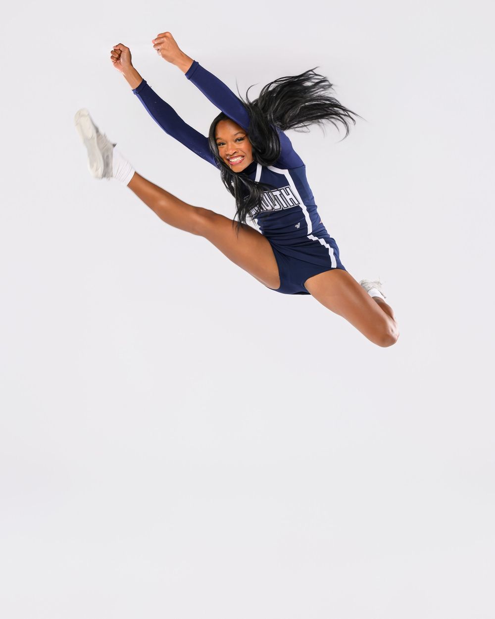 Gymnast in navy blue leotard performs a split jump with graceful arm positions against white background.