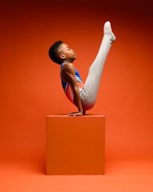 Young gymnast performs impressive V-hold pose on orange cube against vibrant orange background.