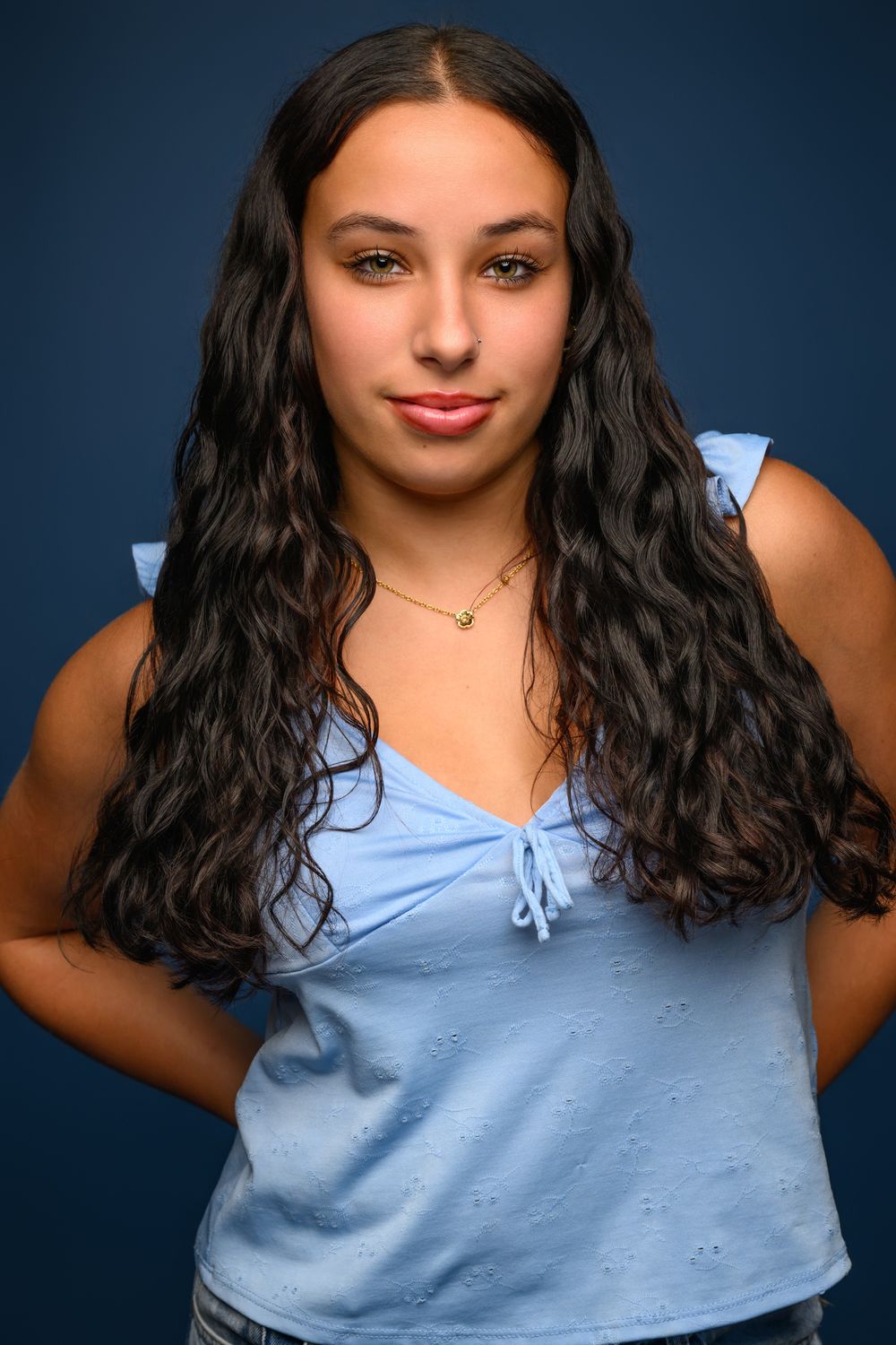 Young woman with long wavy dark hair wearing a light blue sleeveless top, posing against a dark blue background.