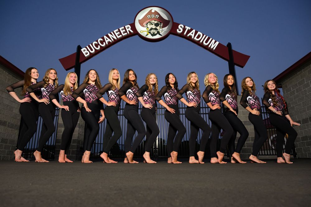 A group of young women in matching uniforms pose in front of Buccaneer Stadium at dusk.