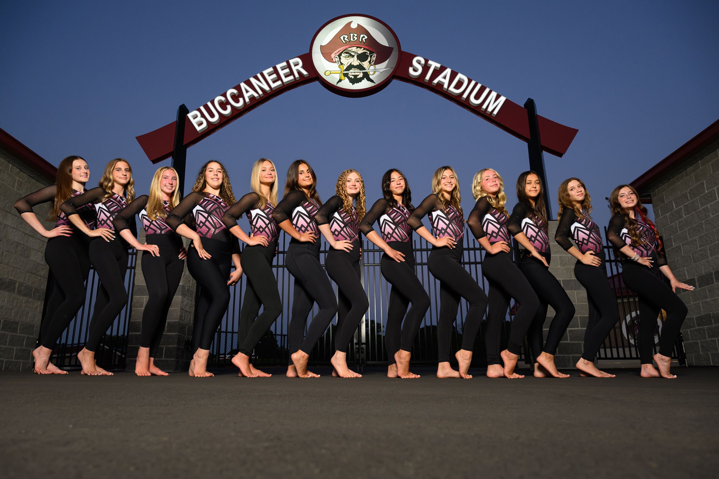 A group of young women in matching uniforms pose in front of Buccaneer Stadium at dusk.