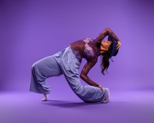 Contemporary dancer in gray pants and burgundy top performs expressive movement against purple backdrop.