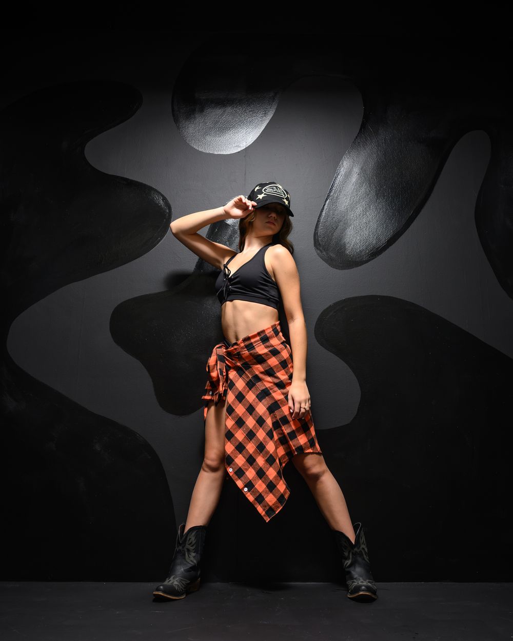 A dancer poses confidently in a black crop top, plaid skirt, and boots against a dark abstract background.