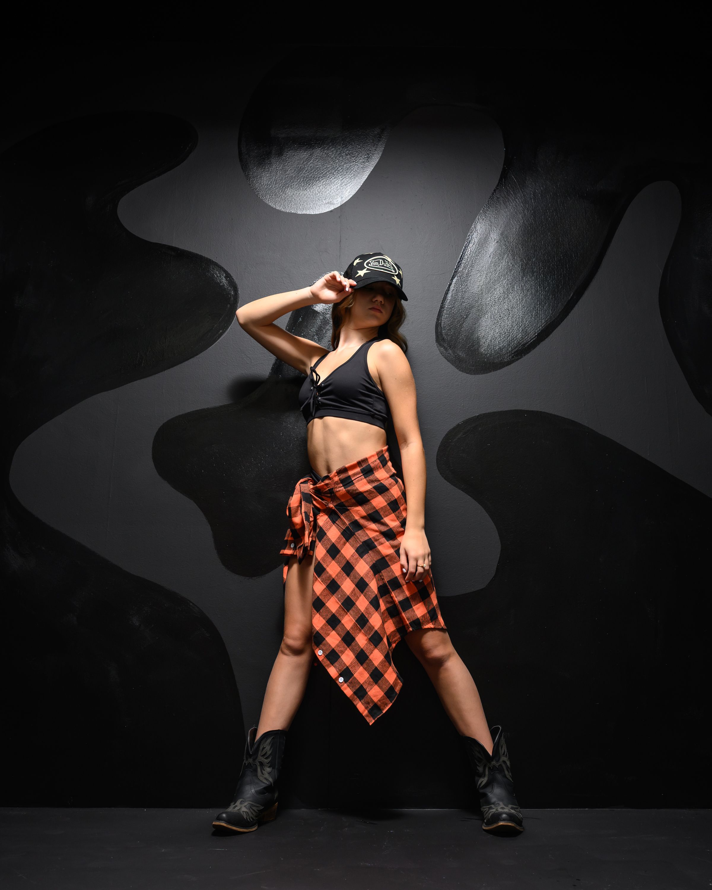 A dancer poses confidently in a black crop top, plaid skirt, and boots against a dark abstract background.