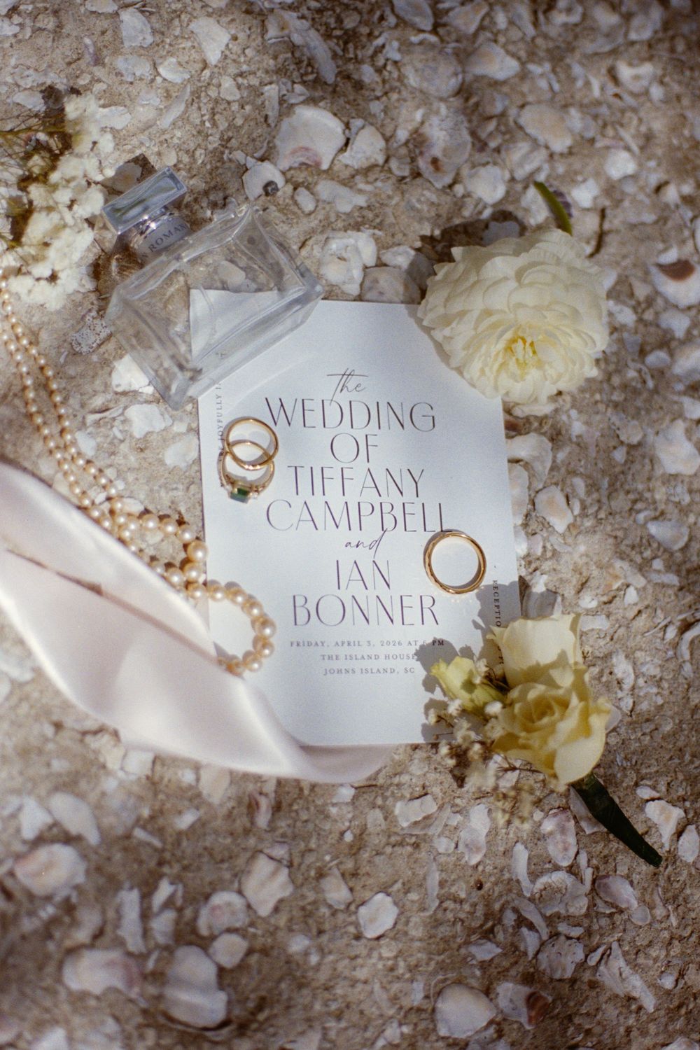 Wedding invitation for Tiffany Campbell and Ian Bonner displayed on sandy beach with gold rings, pearl necklace, and white flowers.