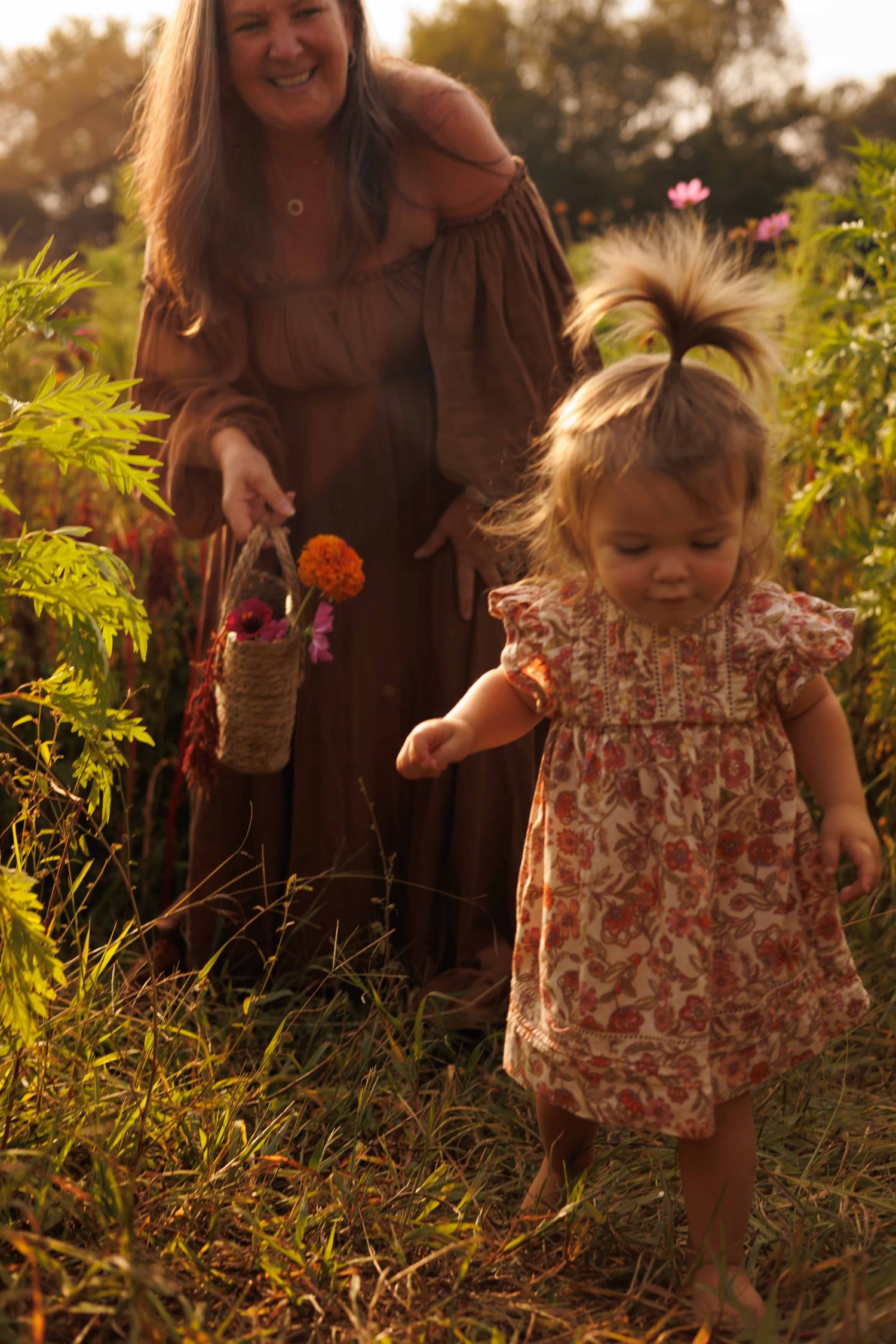 A floral-dressed toddler and someone in brown play together in golden sunlight on grassy ground.