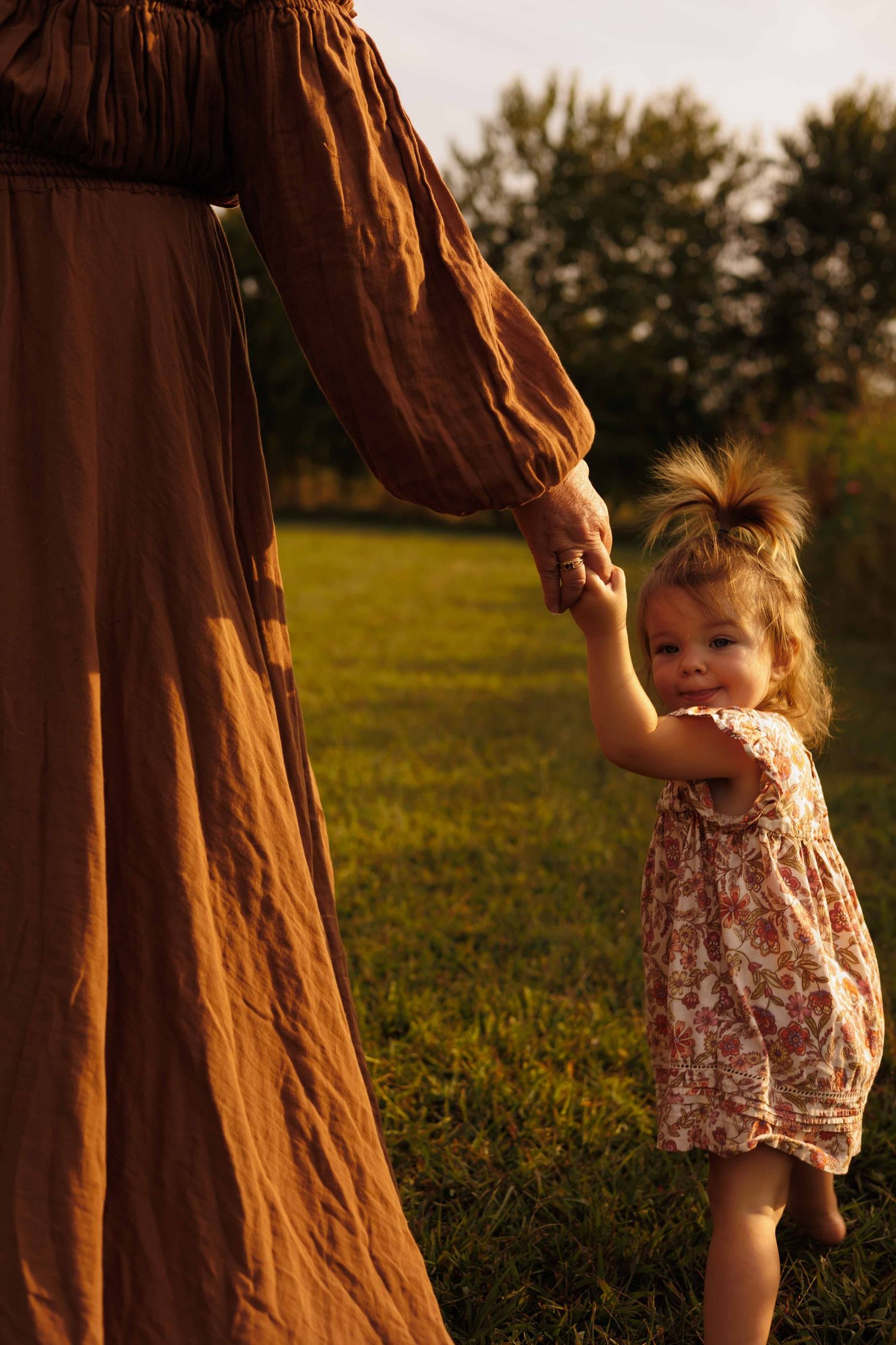 A toddler in a floral outfit runs through grass during golden hour while an adult watches.