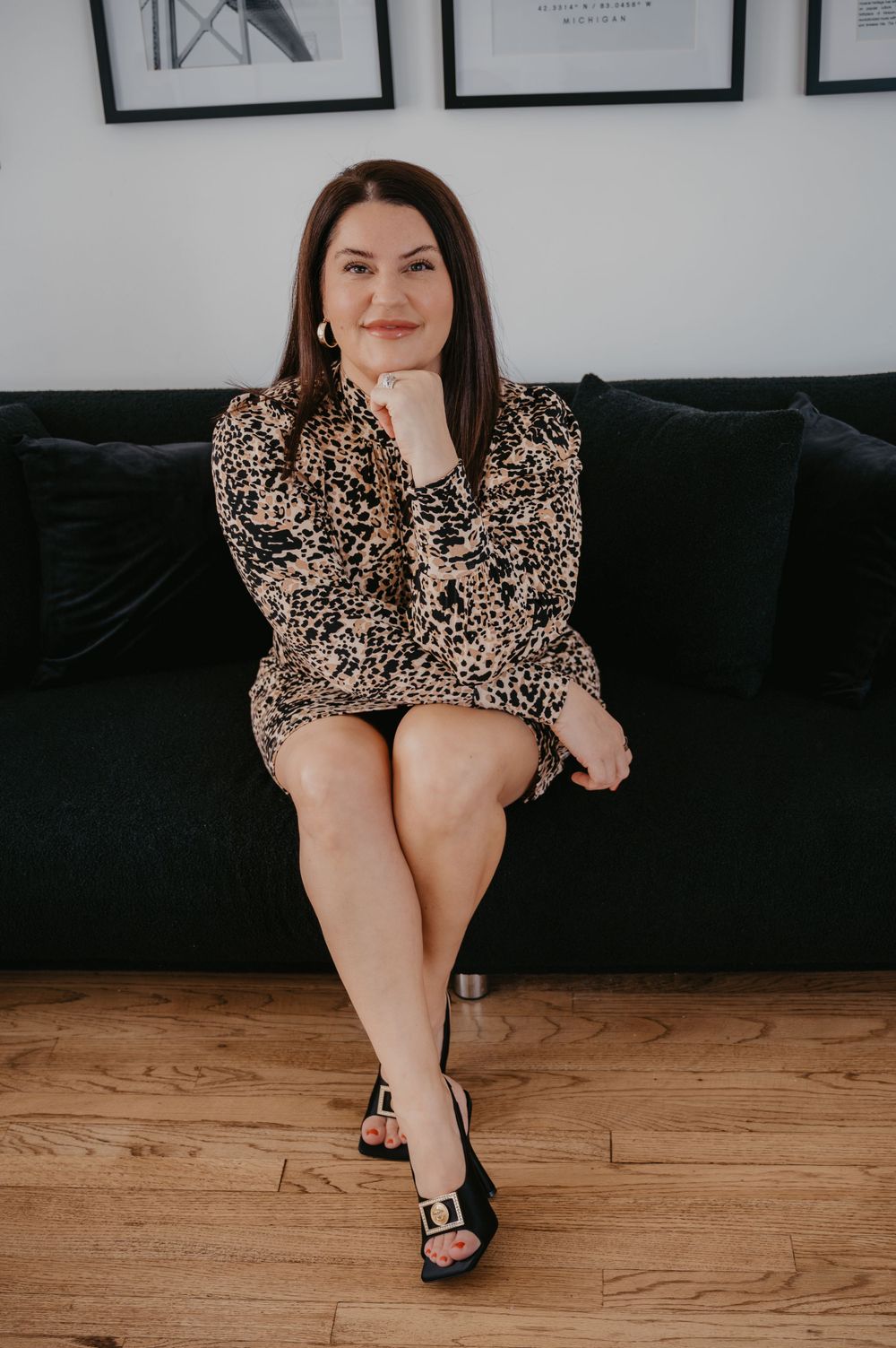 Brunette woman in leopard print mini dress sitting confidently on a black velvet sofa with heels.