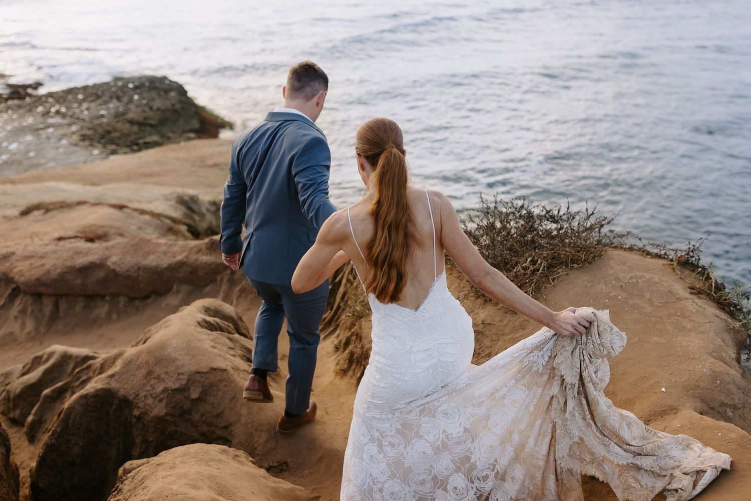 Couple shares adventurous moments on rocky coastal cliffs overlooking the ocean during sunset bridal session.