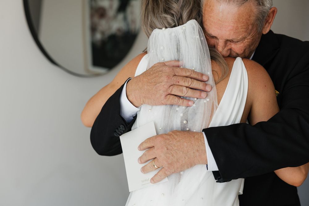 Elderly couple sharing an emotional wedding hug, bride in white gown and groom in black suit, near a round mirror.