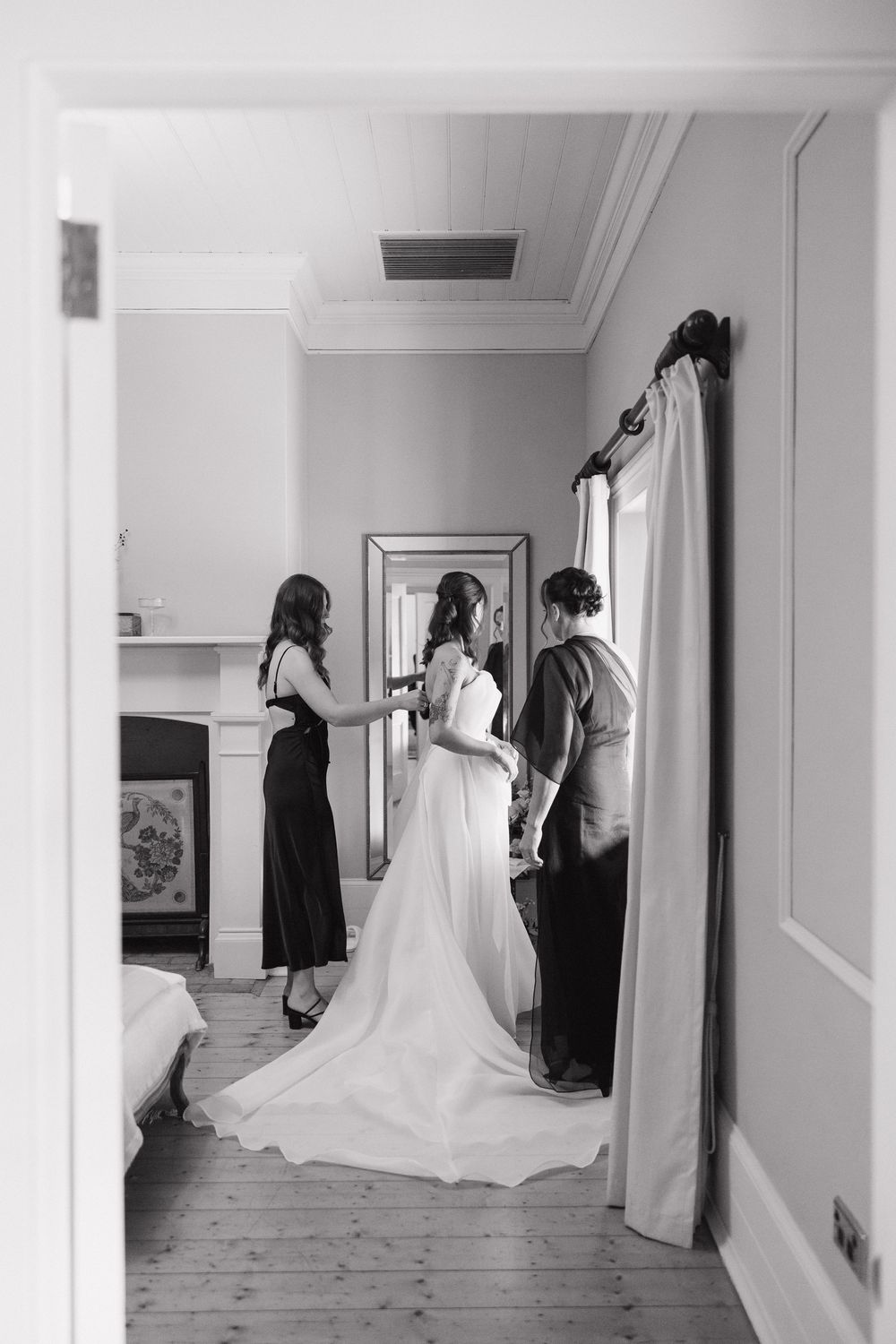 Black and white photo of a bride in a white gown being helped by two bridesmaids in a elegant room.