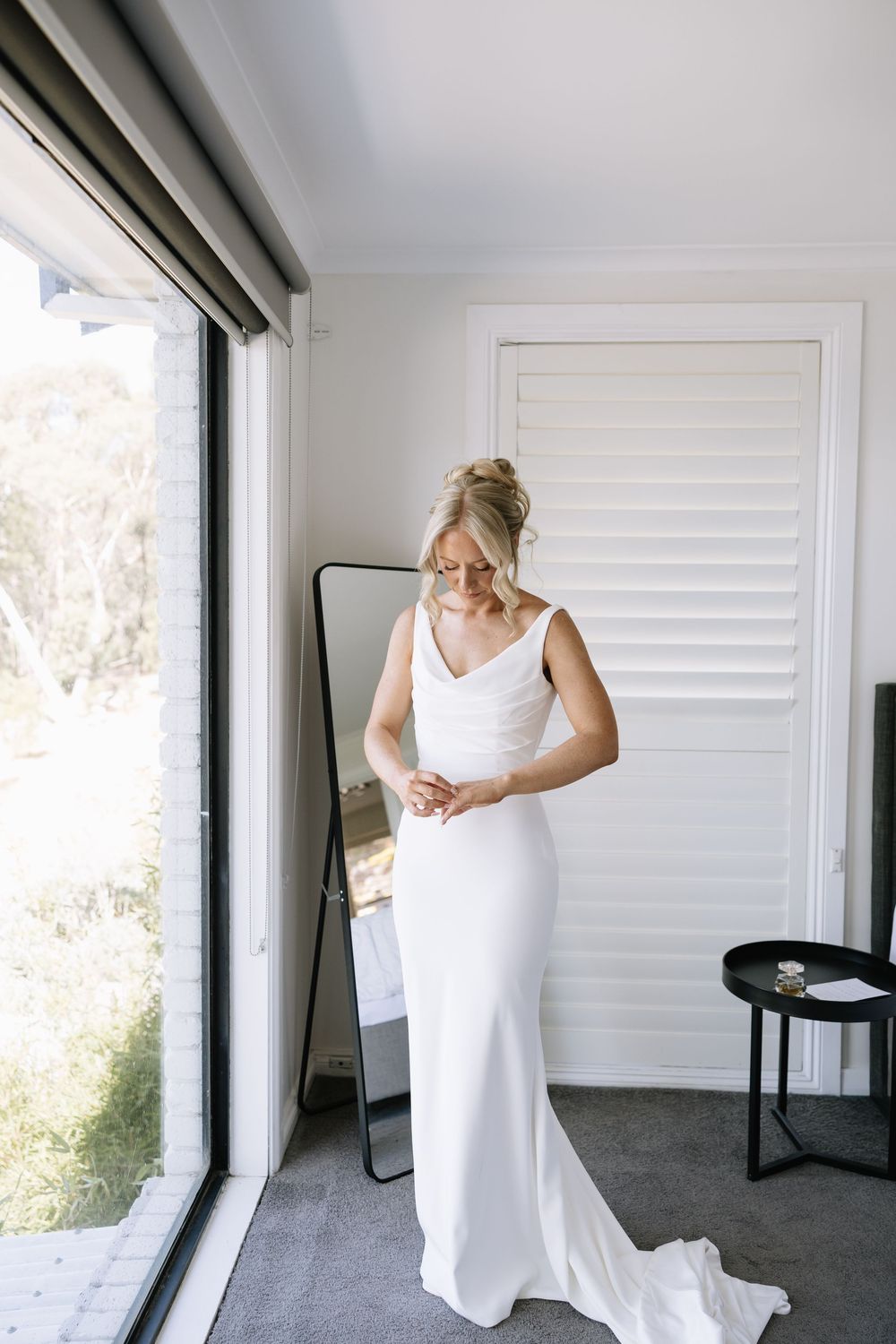 Bride in a sleek white fitted wedding gown adjusting her dress in front of a mirror by a bright window.