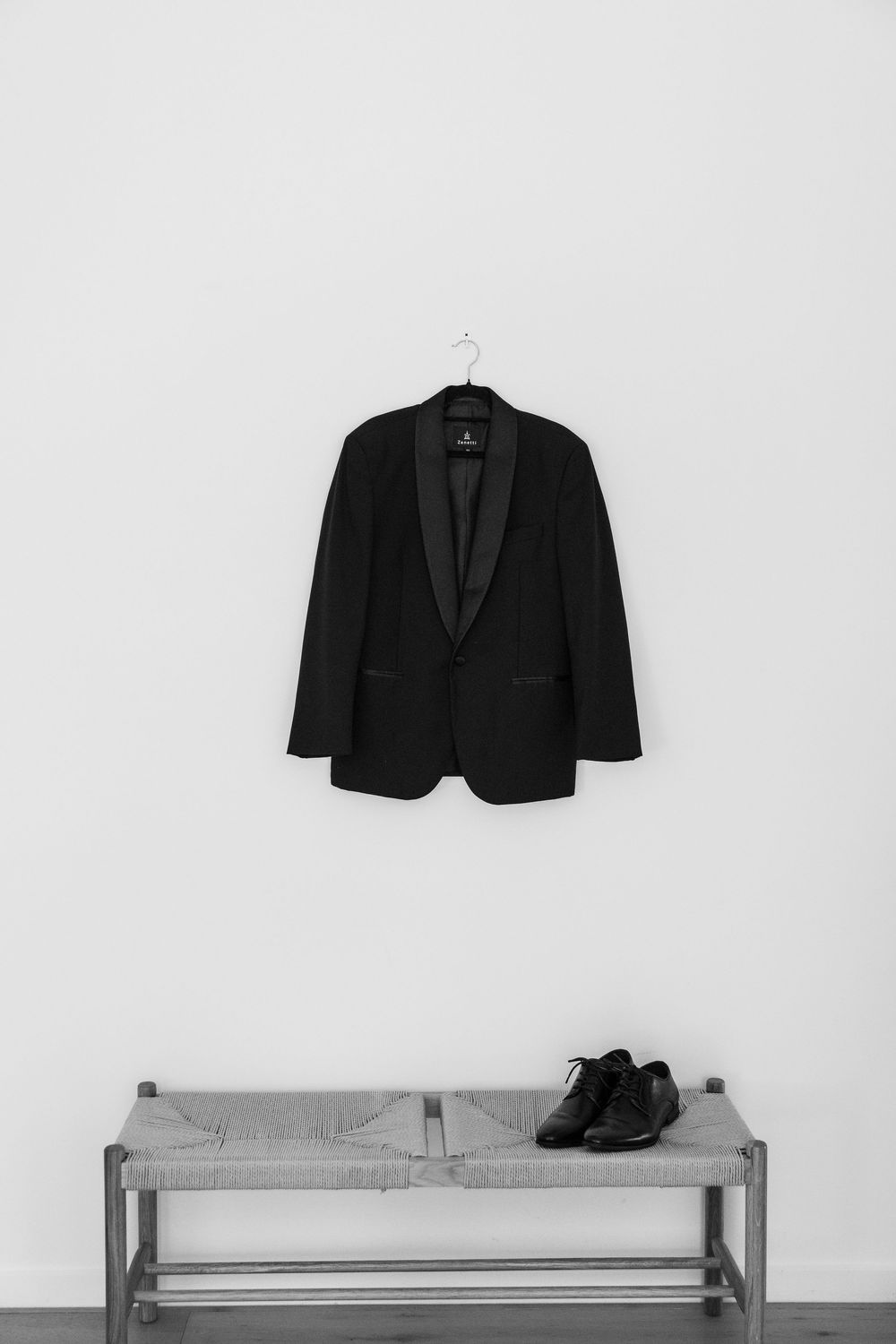 Black tuxedo jacket on hanger above a bench with dress shoes in minimalist monochrome setting.