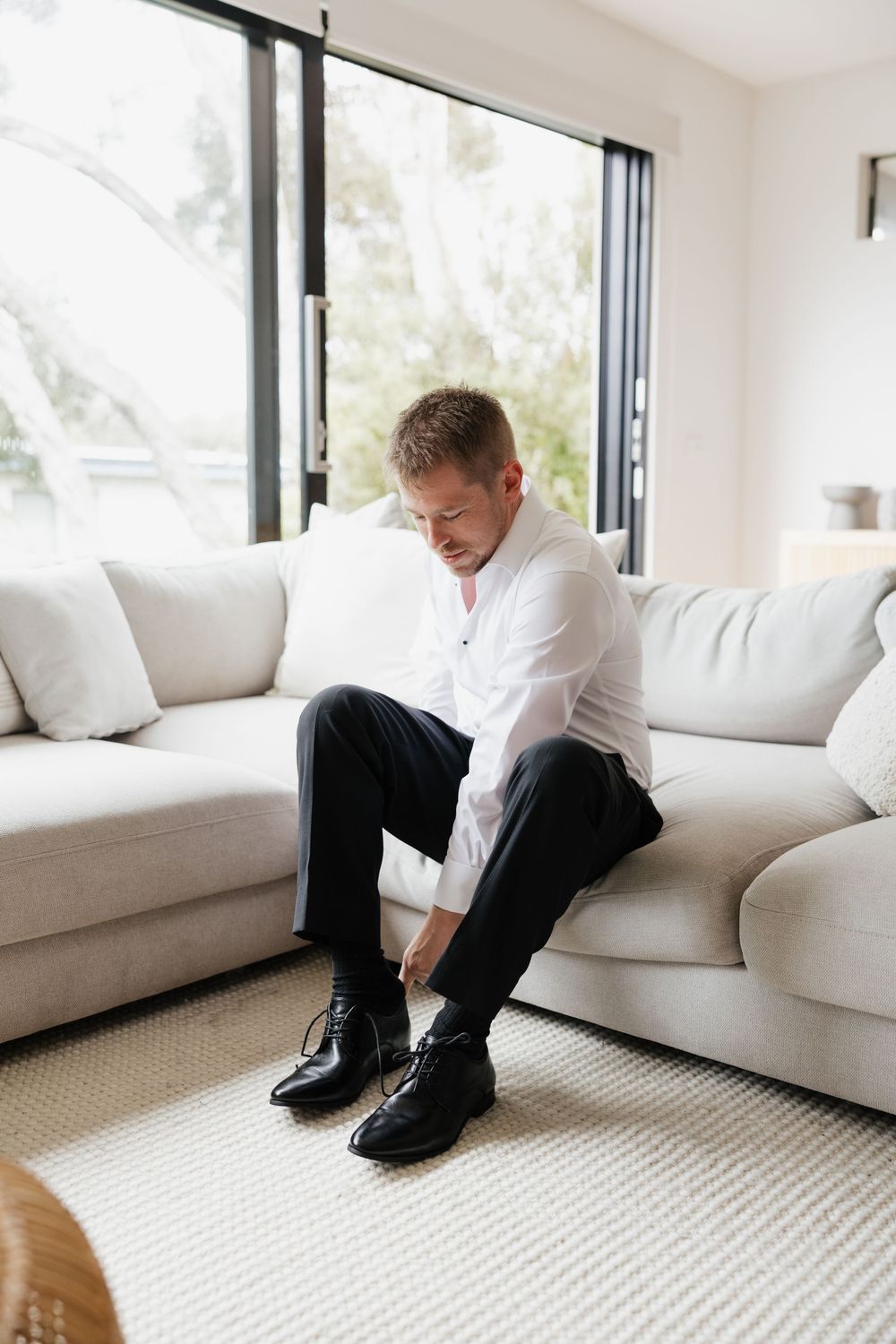 Man in white shirt and black trousers sitting on sofa tying his black dress shoes.