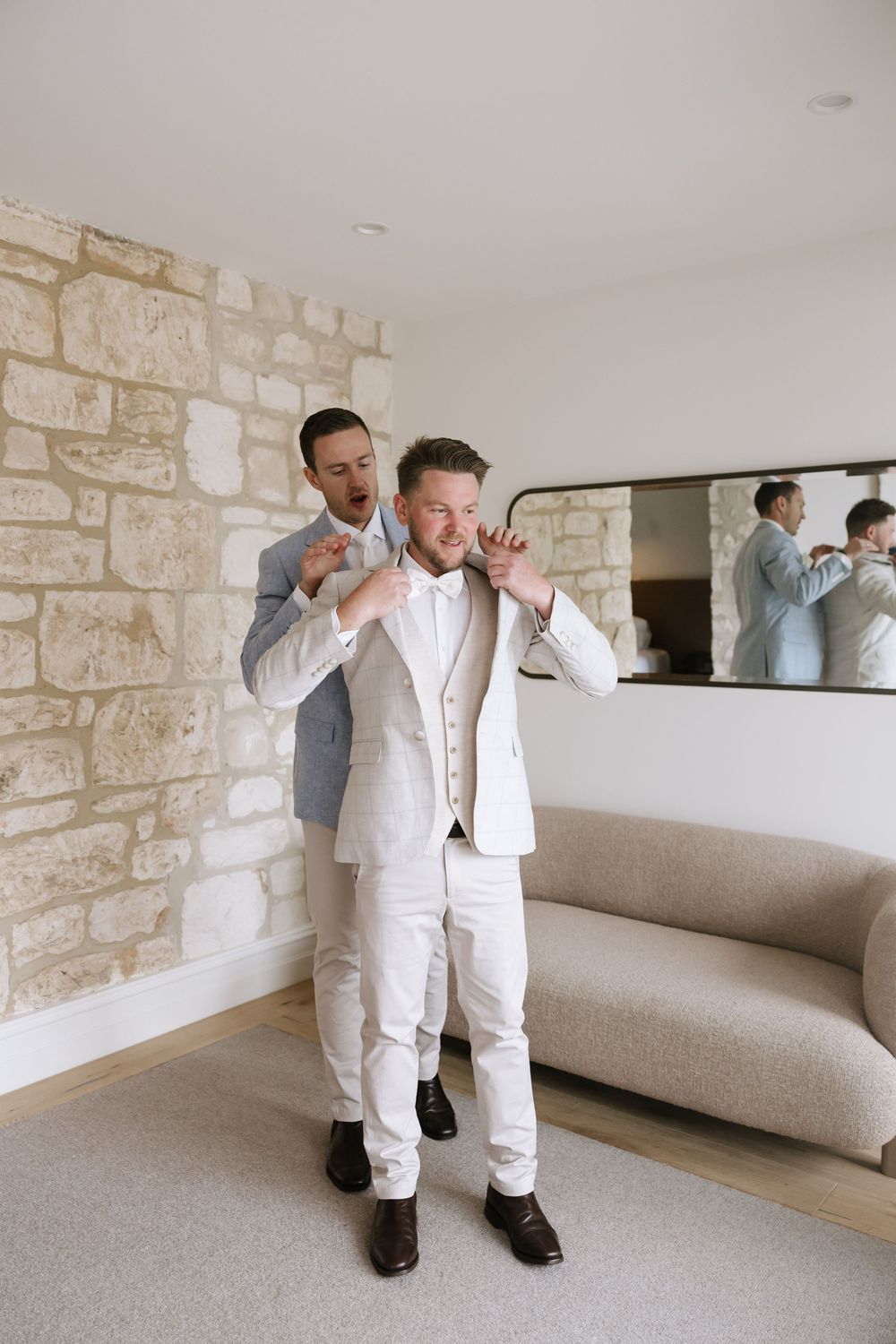 Groom in white suit gets help from groomsman adjusting collar before wedding ceremony in elegant room.