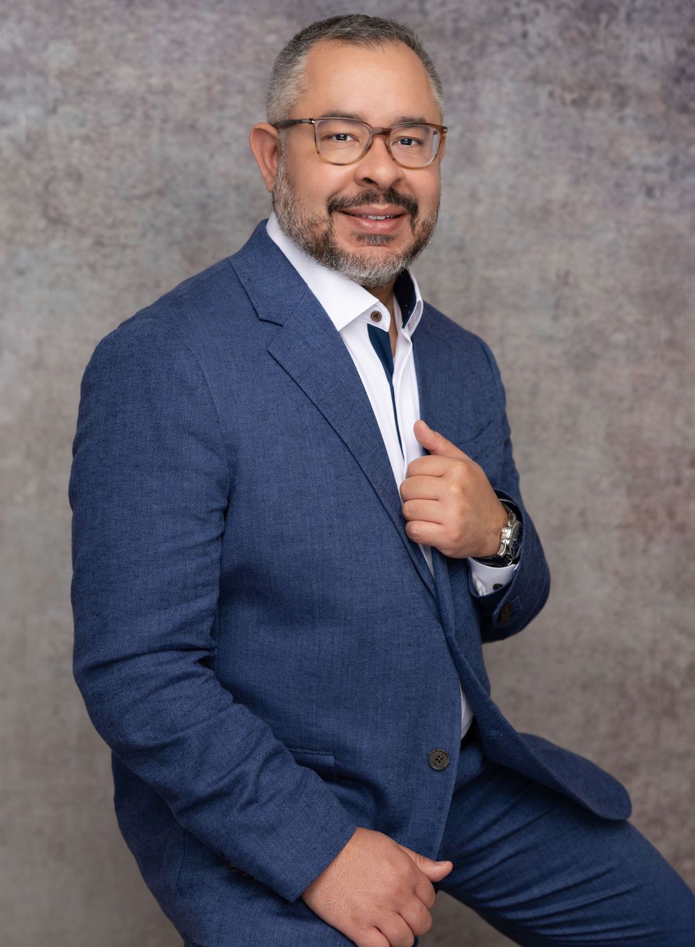 Professional headshot of a person in navy suit and glasses against textured gray background.