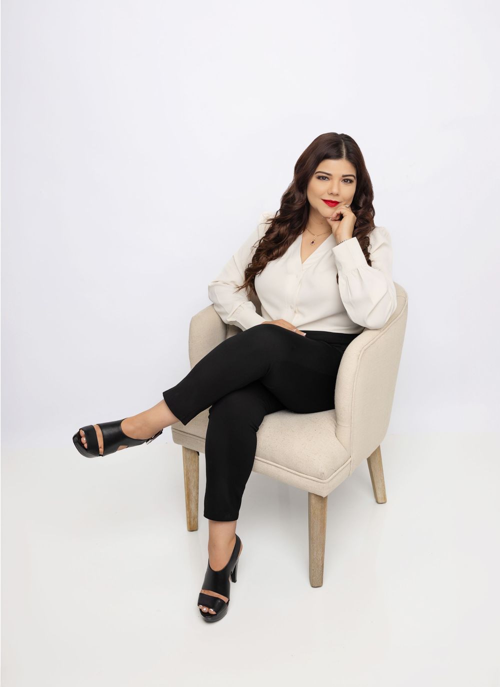 Woman in white blouse and black pants sitting elegantly in beige armchair against white backdrop.