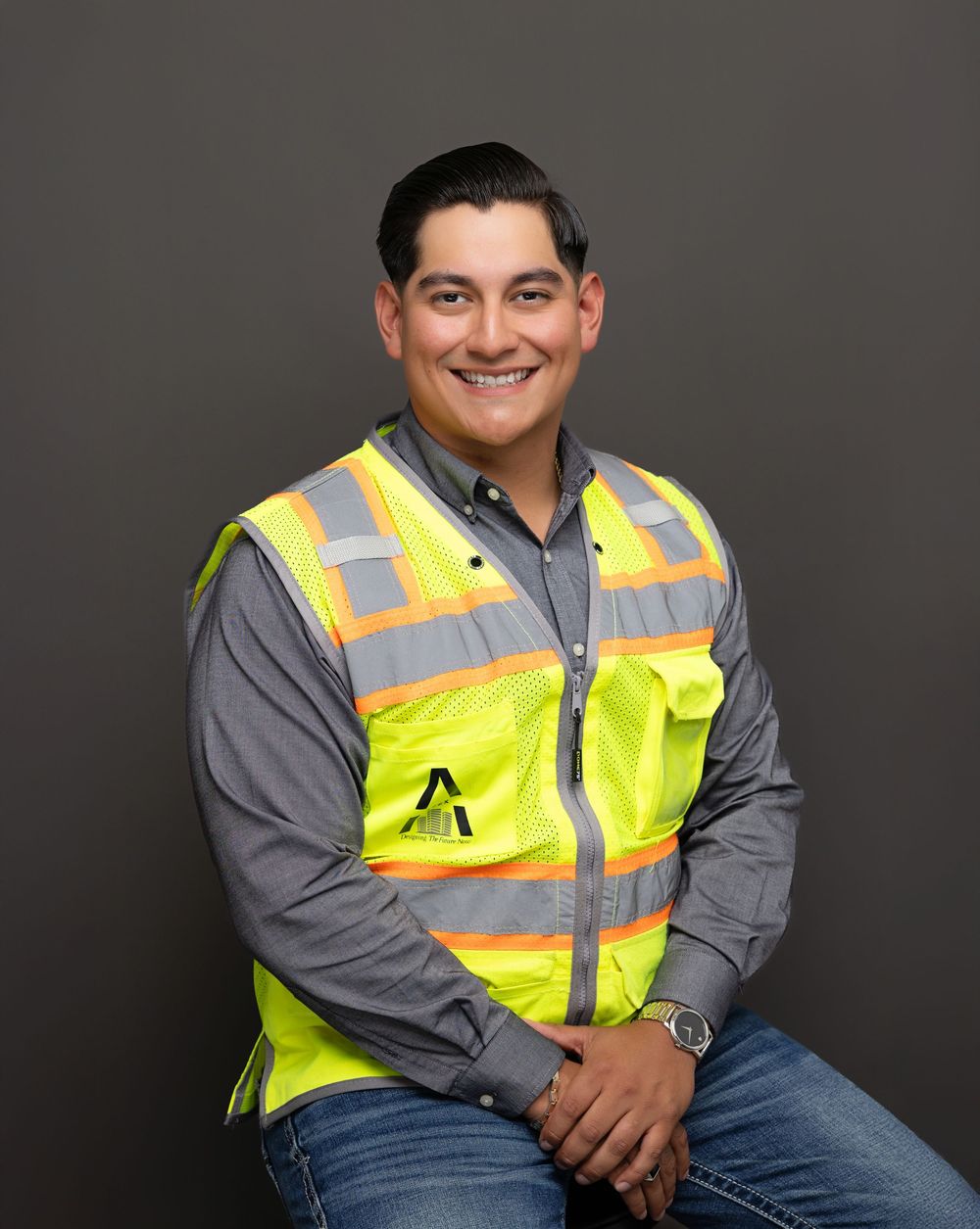 Professional portrait of someone in a reflective yellow safety vest and gray long-sleeve shirt against a dark background.