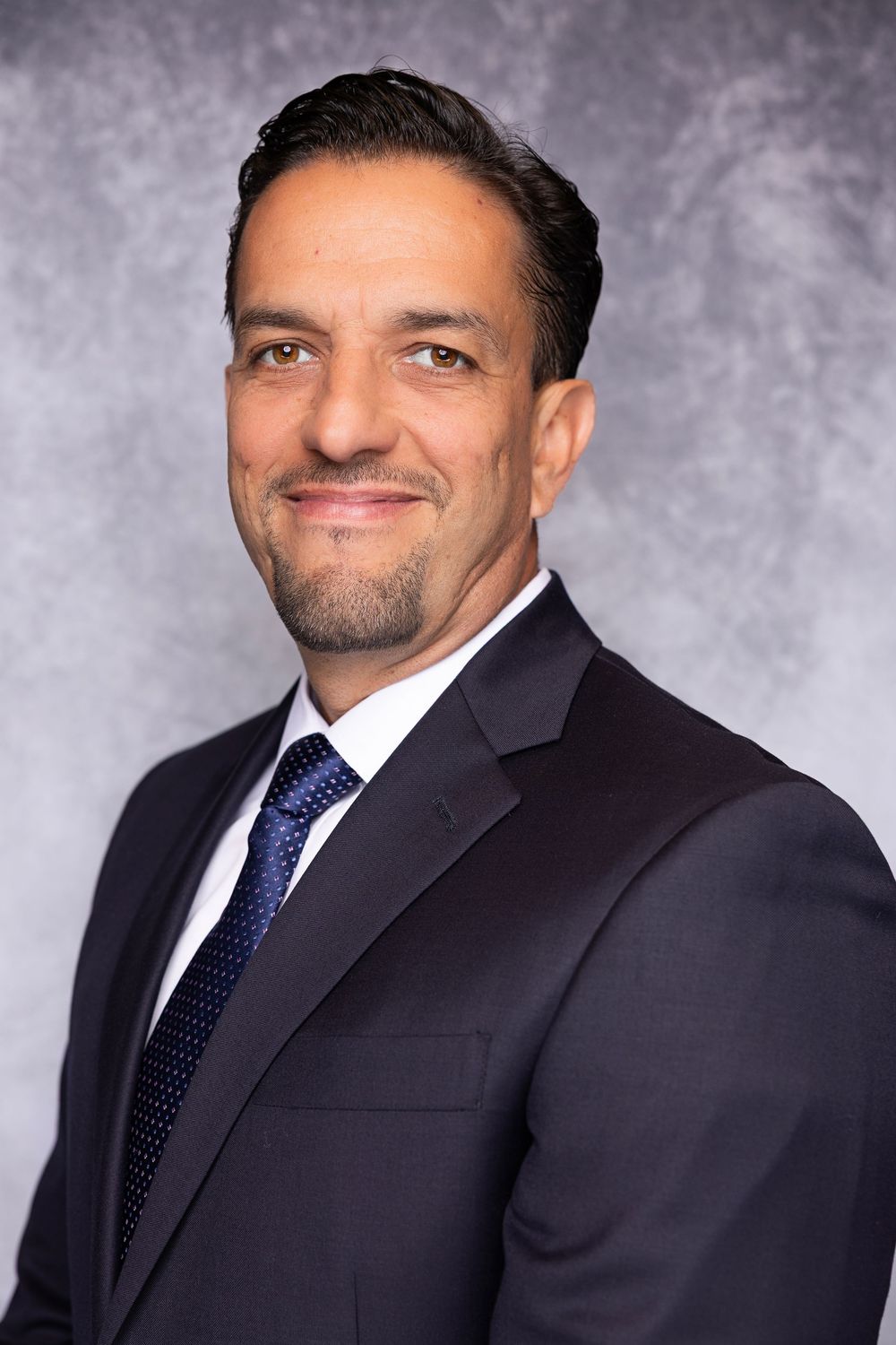 Professional headshot of person in dark suit and blue tie against gray studio backdrop.