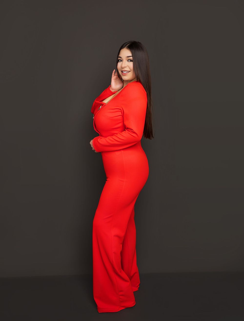 Elegant figure in a form-fitting long red dress poses sideways against a dark backdrop with a bright smile.