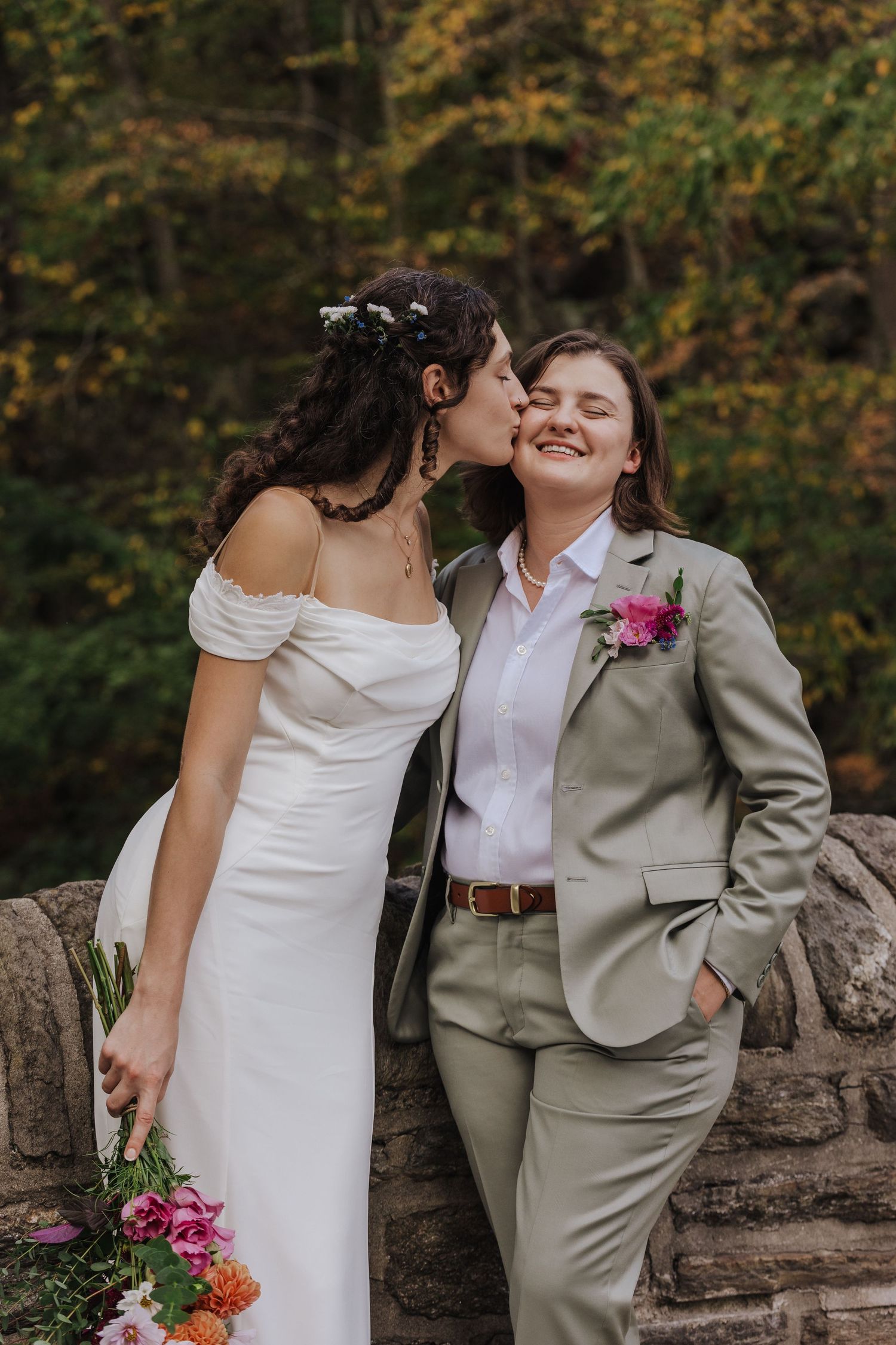 A lesbian couple in wedding attire share an intimate moment against a stone wall in Wissahickon Park with autumn foliage in the background.