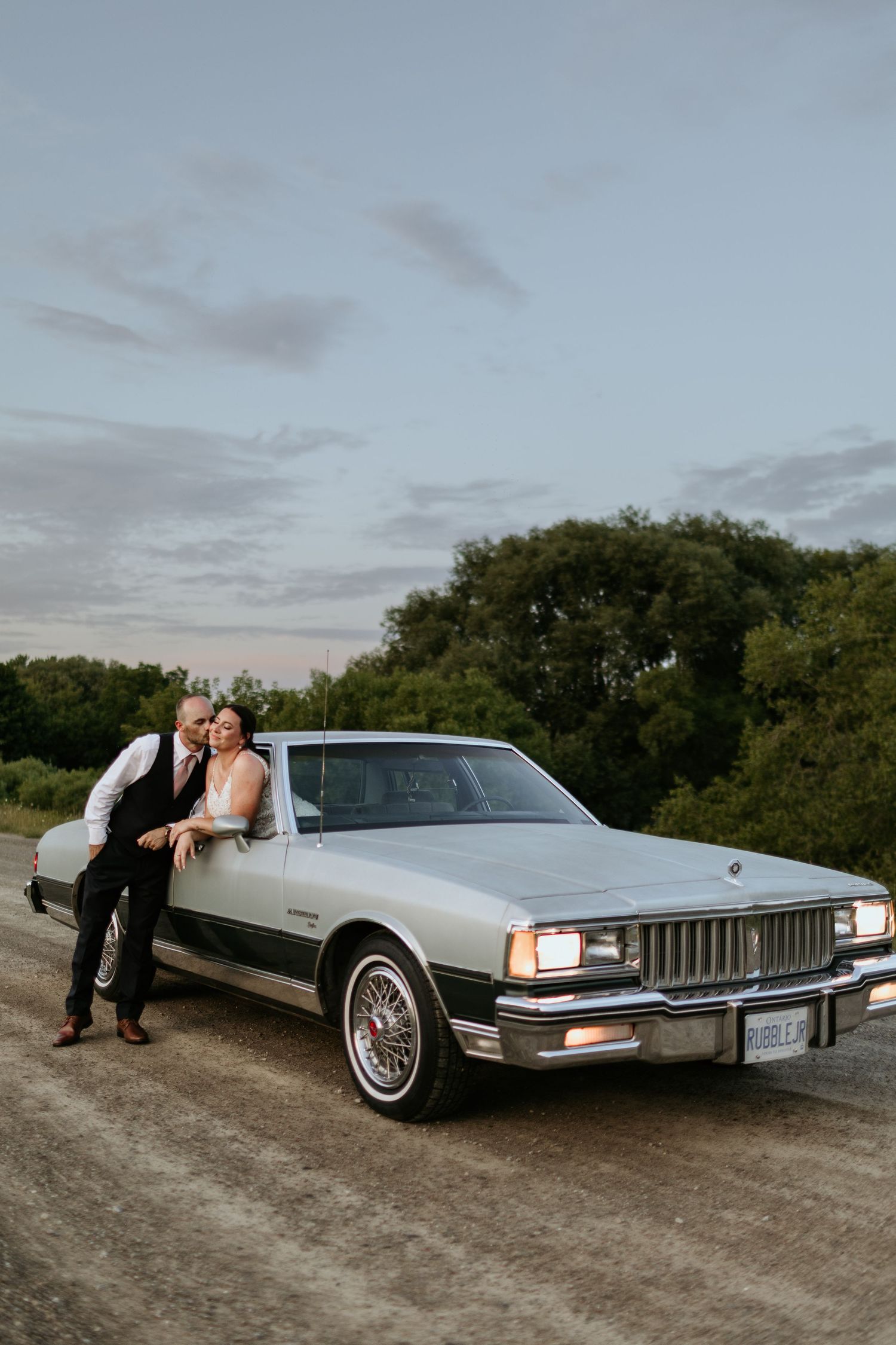 A classic silver Pontiac Caprice parked on a dirt road at dusk with a couple posing beside it.