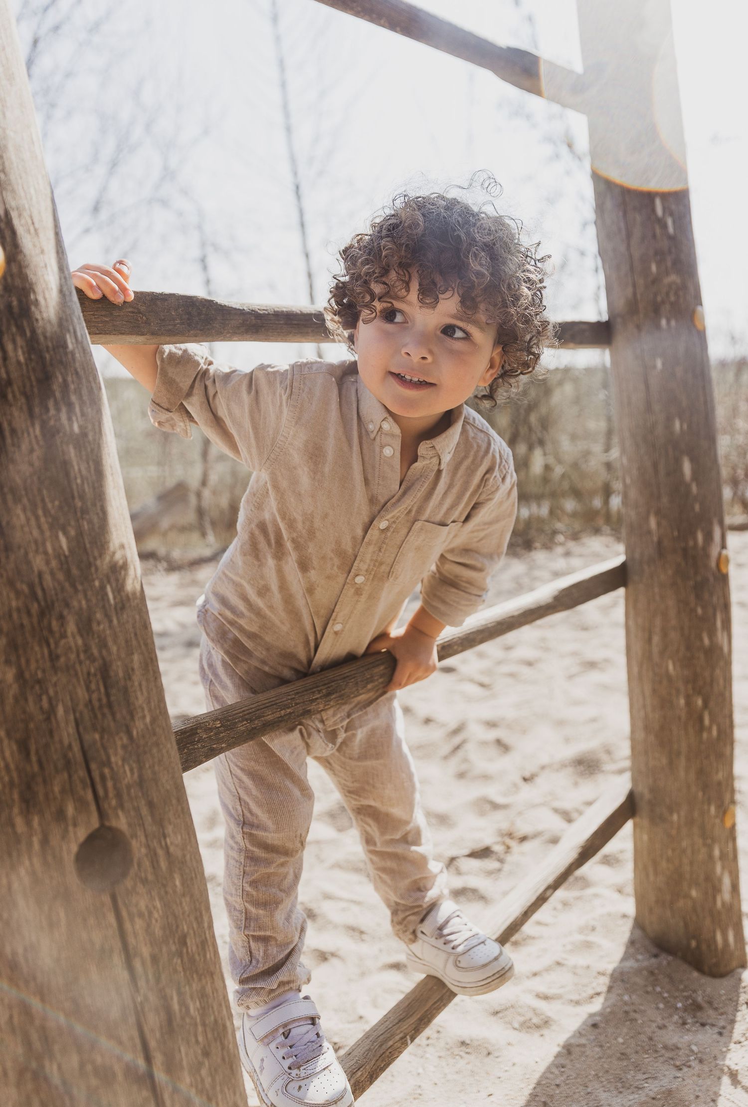 Young child wearing beige outfit climbing on wooden playground structure at beach. campagne shoot. kinderkledingfotografie.