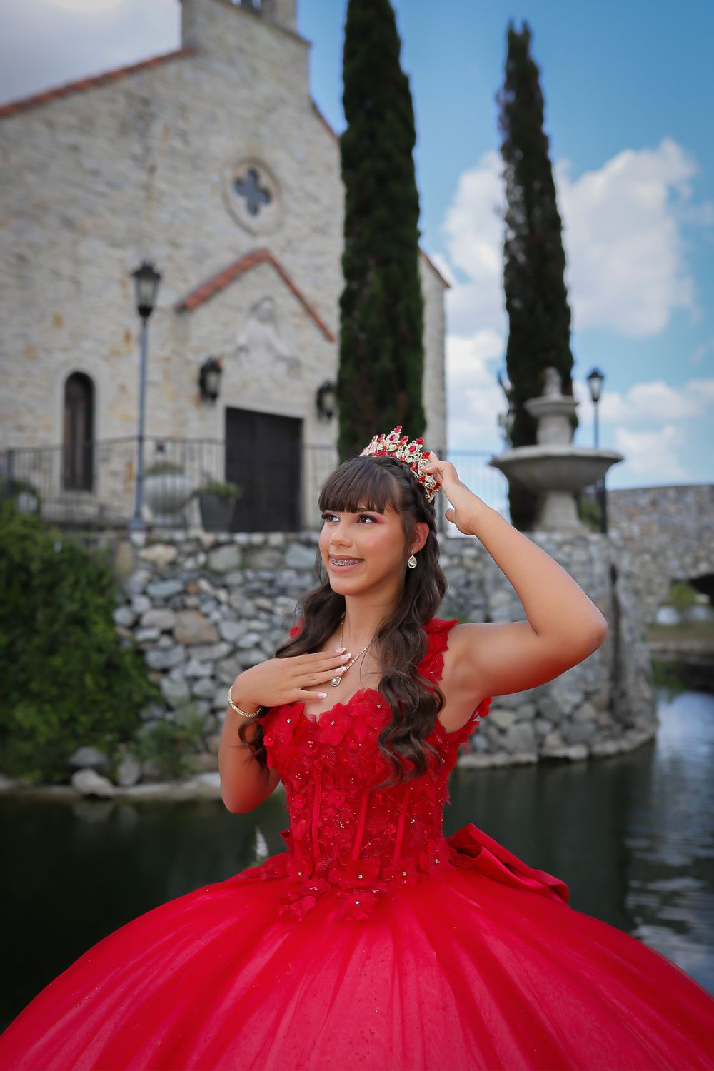 A red quinceañera dress is showcased in front of a white stone church with tall cypress trees and lamp posts.