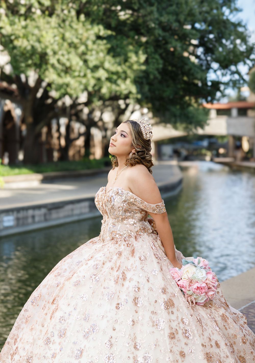 A person in a blush pink quinceanera gown with floral details poses along a waterway holding a pink bouquet.
