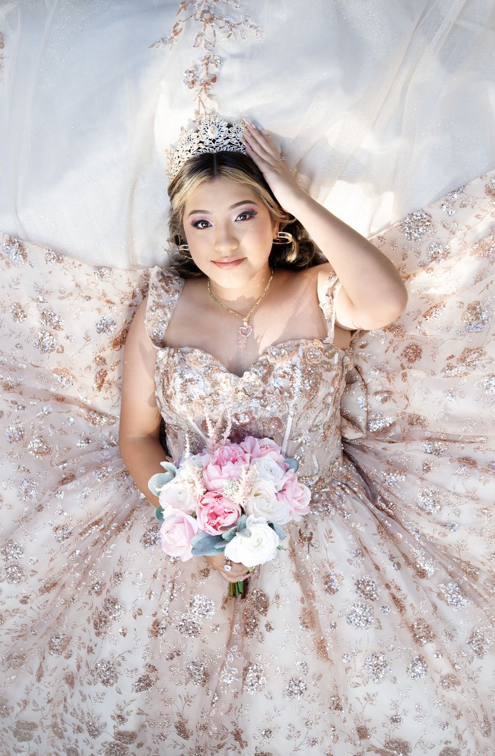 A person wearing a rose gold sequined ball gown with tiara holds a pink and white floral bouquet while lying on white fabric.