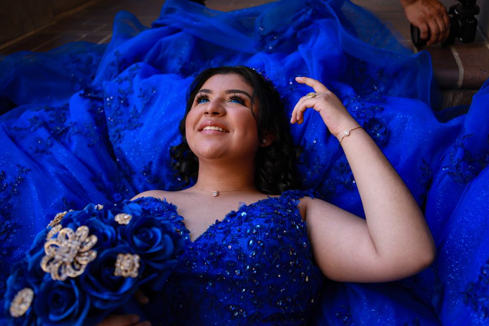 Someone wearing a sparkling royal blue quinceañera dress lies on the floor with their dress elegantly spread out around them.