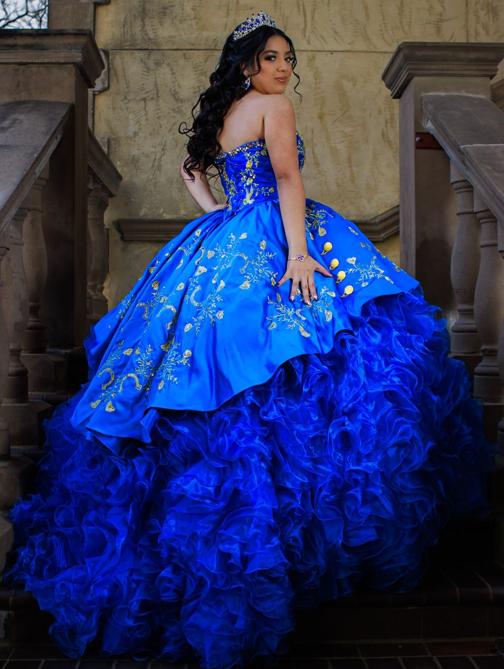 Elegant royal blue quinceanera ball gown with intricate embroidery and ruffled organza skirt photographed against stone wall.