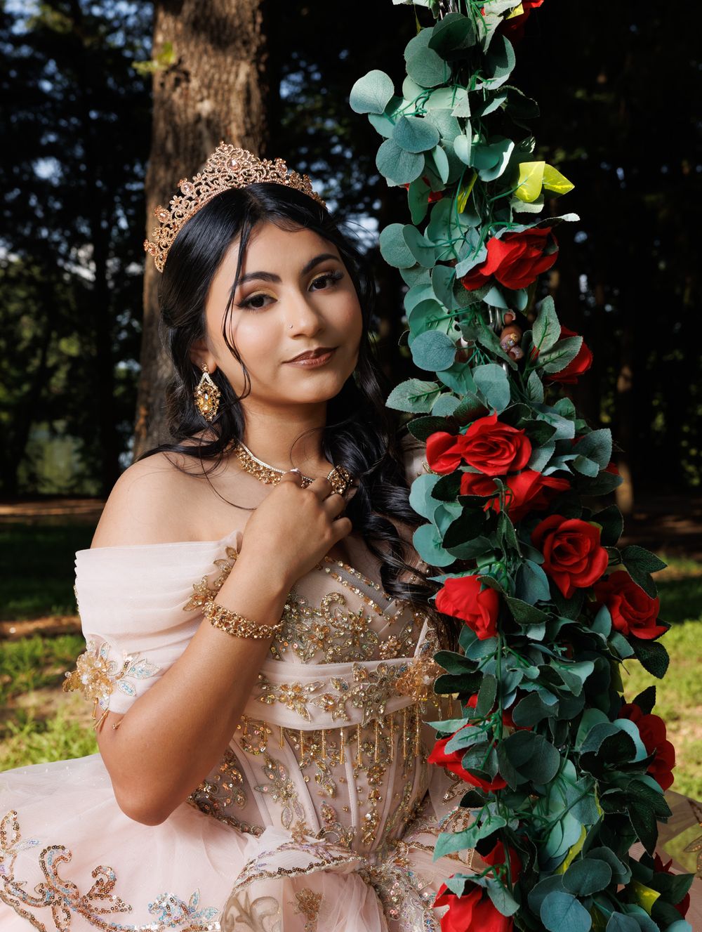 Ethereal bridal portrait with blush gown and red roses on a floral arch in outdoor garden setting.