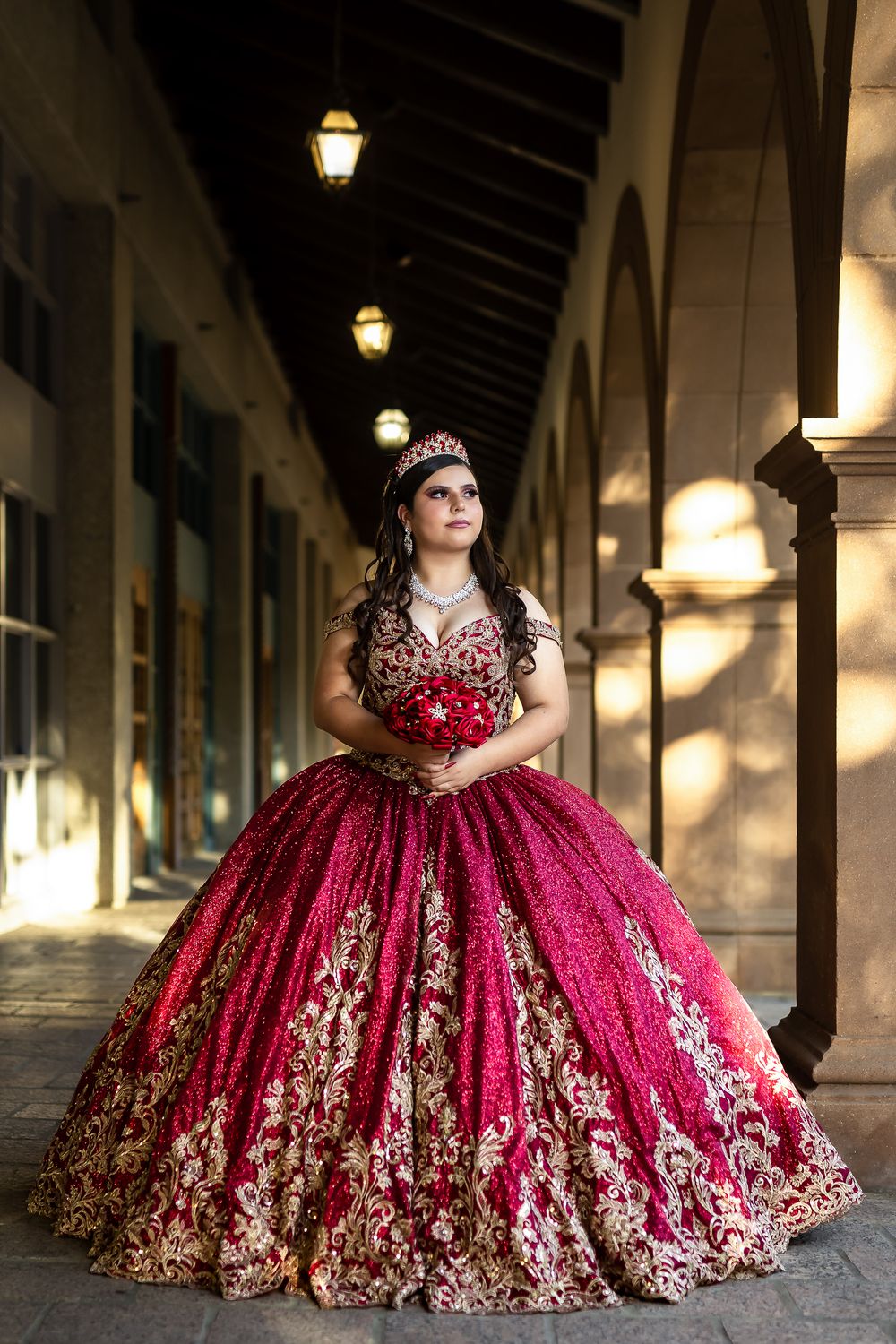 A person in an ornate burgundy and gold quinceañera ball gown stands in a covered outdoor walkway with arches and lanterns.