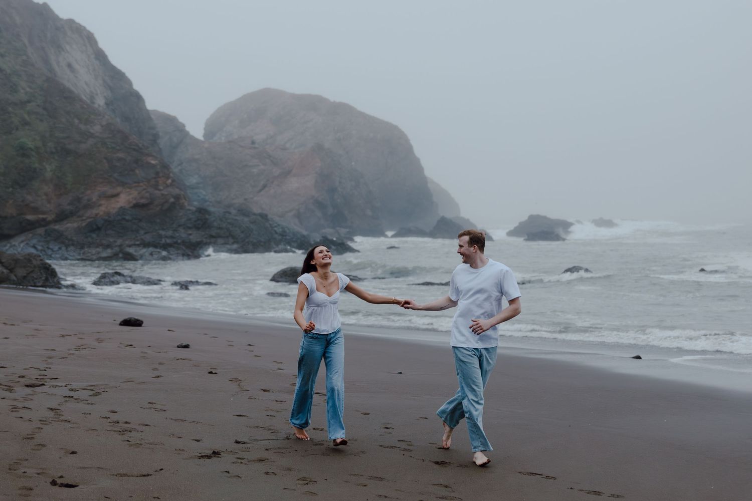 Two people dance playfully together on a misty beach with rocky cliffs in the background.