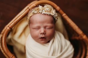 A sleeping newborn baby wrapped in white fabric lies peacefully in a woven basket wearing a delicate gold headpiece.