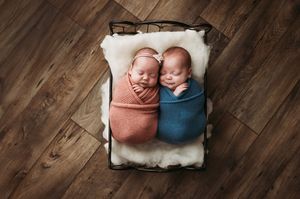 Two sleeping newborn babies swaddled in rust and navy colored wraps lie side by side on a wooden floor backdrop.