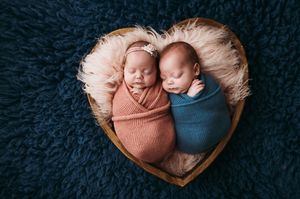Two swaddled newborn babies sleeping together in a heart-shaped basket on a dark blue textured background.