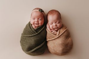 Two newborn babies wrapped snugly in olive green and tan swaddle blankets lying side by side on beige backdrop.